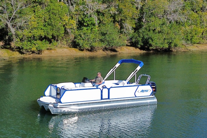 A person steers a small white and blue pontoon boat on a calm lake with tree-lined shore.