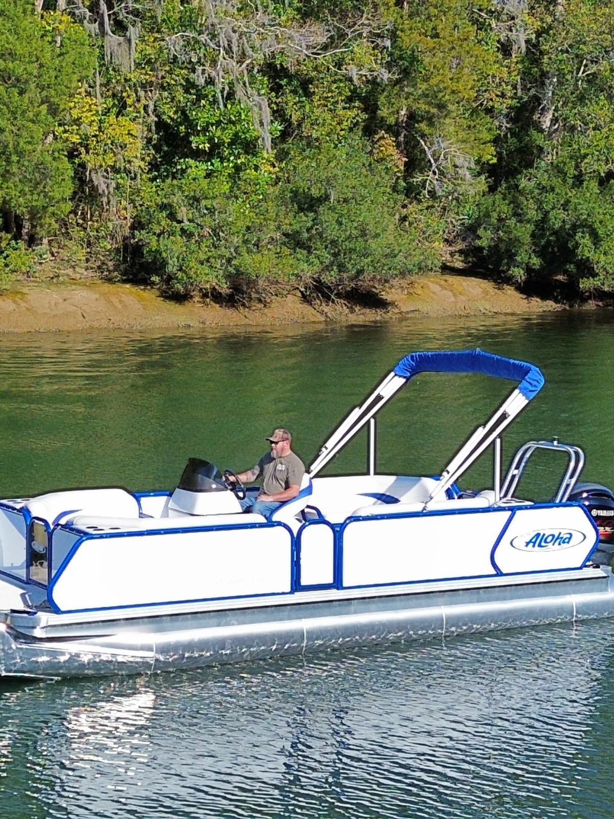 A person steers a small white and blue pontoon boat on a calm lake with tree-lined shore.